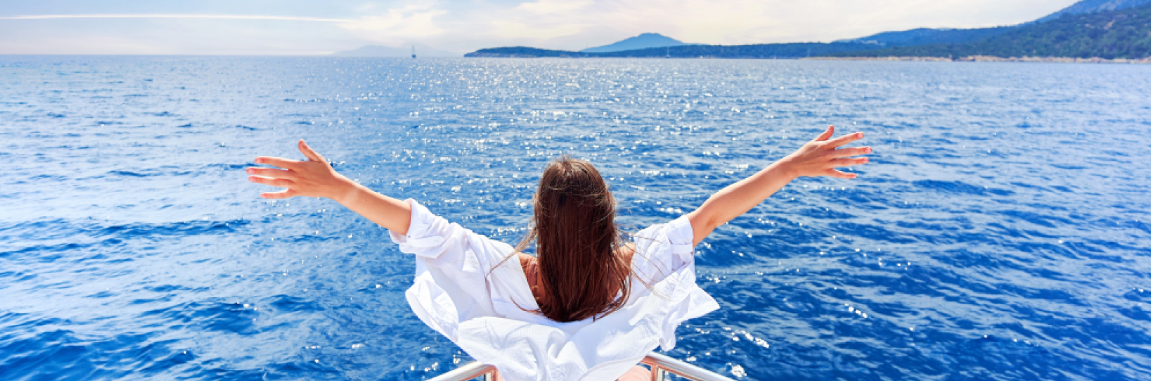 A woman with long brown hair wearing a white off-the-shoulder top stands at the edge of a boat with her arms outstretched, embracing the open sea.This image represents the freedom and joy of travelling with fibromyalgia, highlighting the importance of relaxation, scenic journeys, and mindful travel choices to manage symptoms while exploring the world.
